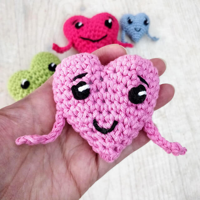 Someone holding a pink crochet heart with arms eyes and a mouth with other coloured hearts in the background