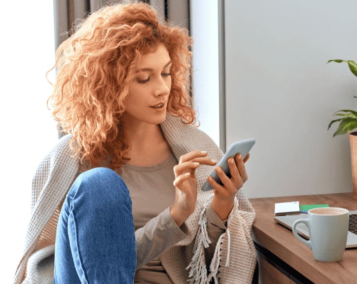 Woman sitting by a desk with a laptop looking at her phone
