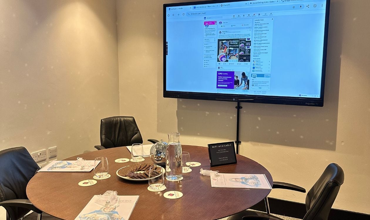 A small meeting room set up for a LinkedIn business training session. Four chairs surround a round table with workbooks, glasses, a jug of water, a plate of biscuits, and a disco ball in the centre. On the wall, a large screen displays a LinkedIn profile, and a sign on the table provides WiFi details. The scene is welcoming and ready for a LinkedIn course for business owners.