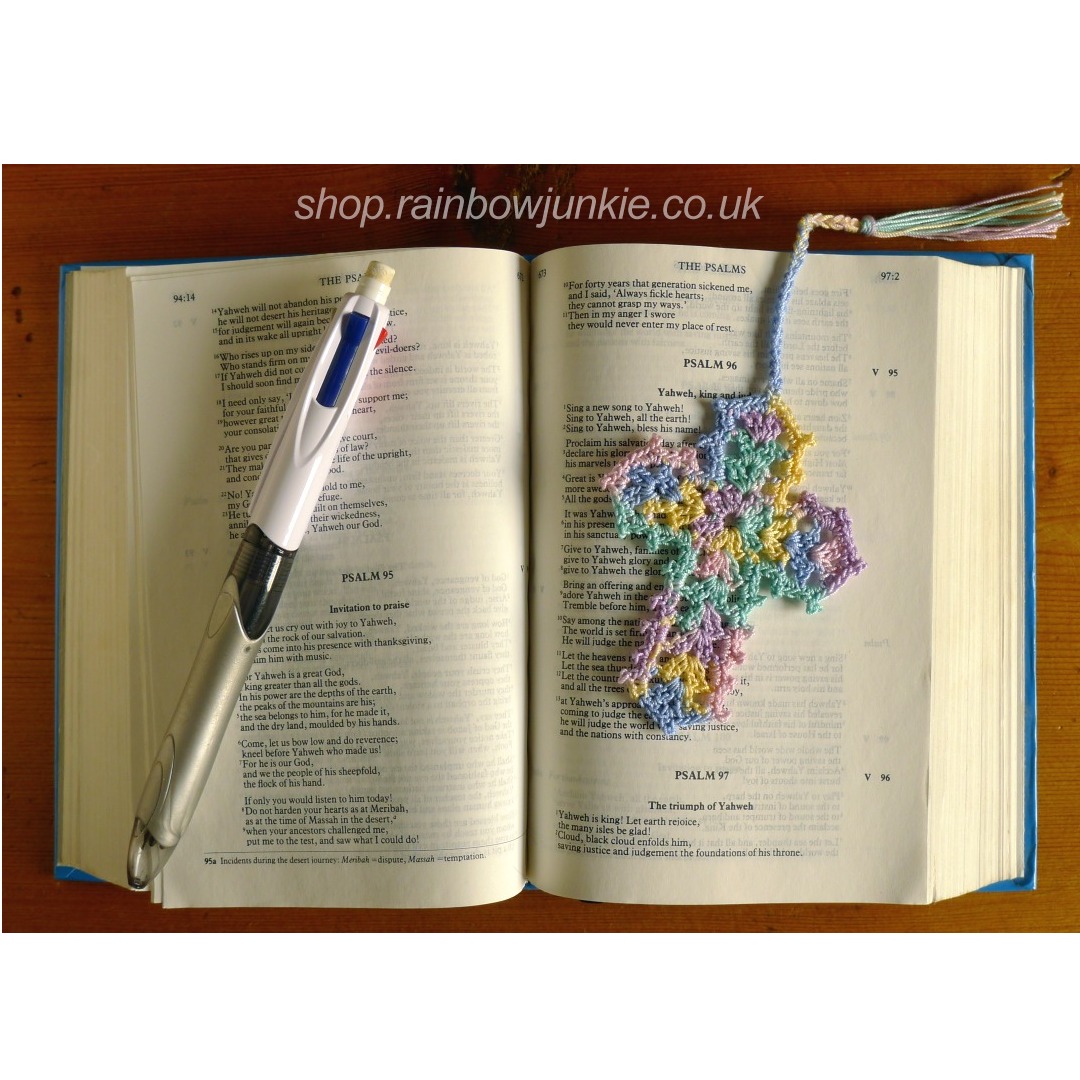 Dainty cross bookmark made in a multicolour cotton thread lying on an open small bible with a ball point pen for size comparison.