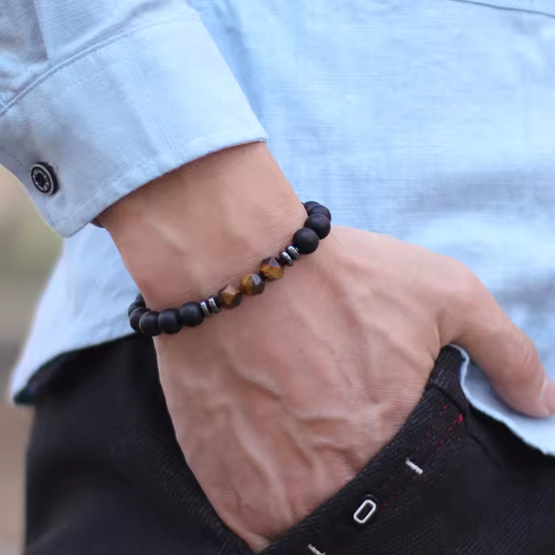 Man wearing a matte black onyx and golden-brown Tiger Eye bead bracelet, featuring faceted central stones and hematite spacers, styled with a light blue shirt and dark trousers.