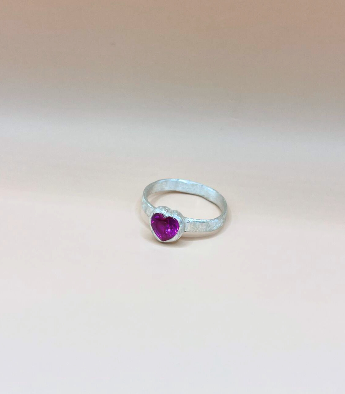 Silver ring with a textured band featuring a heart-shaped pink ruby in a bezel setting against a neutral beige background.