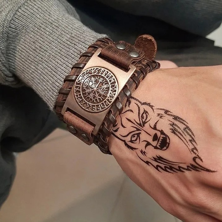 Lifestyle image: Man wearing a wide brown leather Viking cuff bracelet featuring a bronze-tone Vegvisir (Viking compass) and runic emblem. The Norse pagan aesthetic is enhanced by a prominent black wolf head tattoo on his hand and a grey textured sweater.