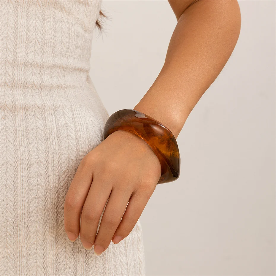 Lifestyle image of a woman wearing a chunky wide cuff bangle made of polished, dark brown tortoiseshell resin. The statement bracelet's unique organic and irregular shape is shown on her wrist, styled elegantly with a textured white dress.