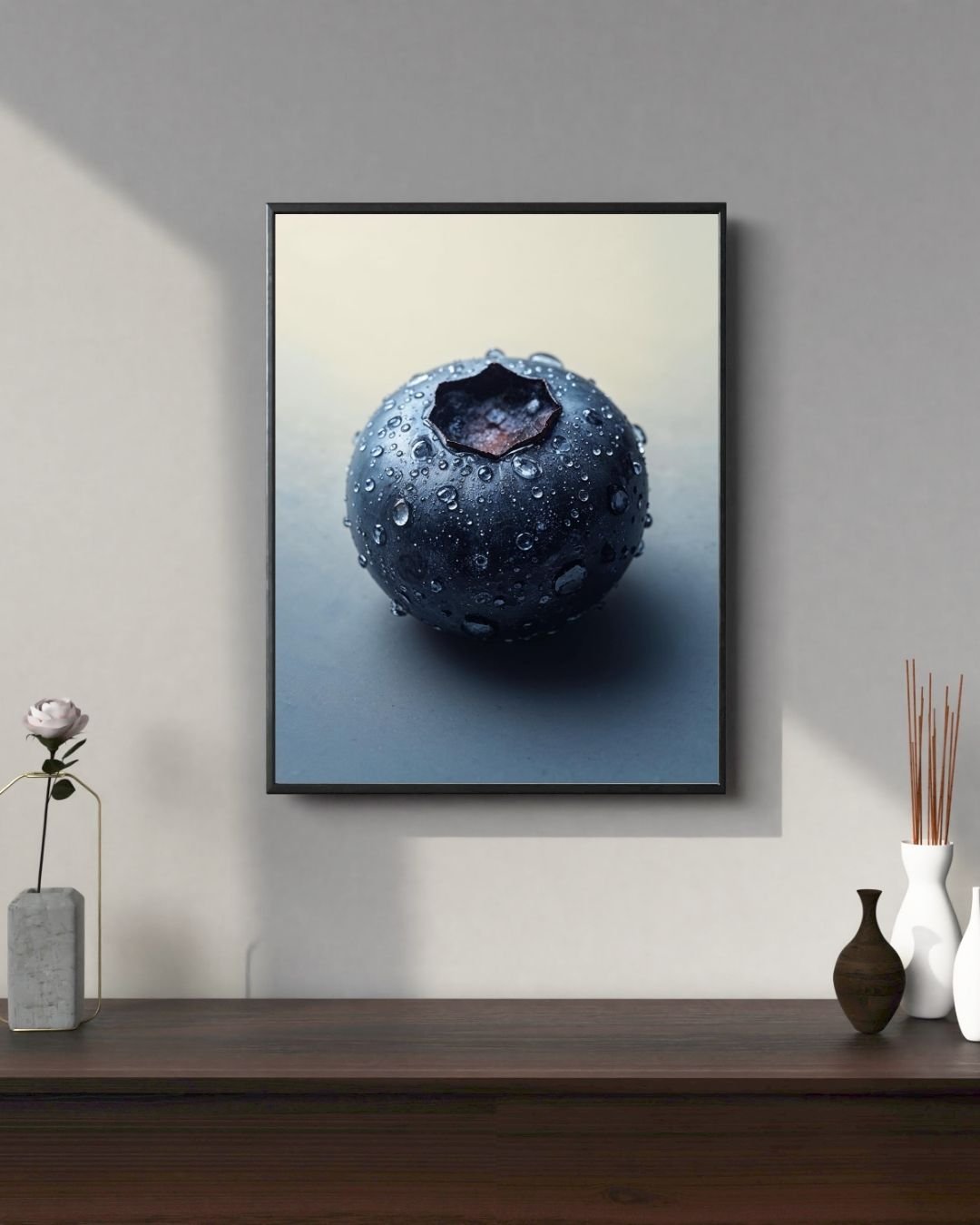 A close-up photograph of a single fresh blueberry covered in small water droplets. The berry has a smooth, dark blue surface with a matte sheen and a star-shaped calyx at the top. The soft lighting and neutral background highlight the texture and moisture