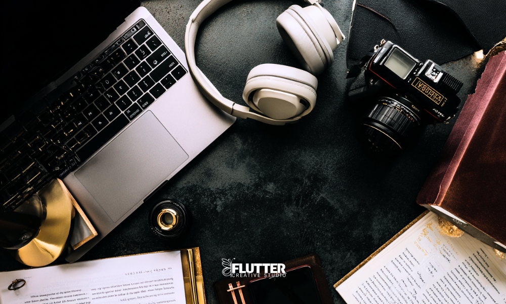 A flat lay image of a creator's desk with a laptop, headphones, a vintage camera, open notebook, and pen, symbolizing digital content creation and editorial work.