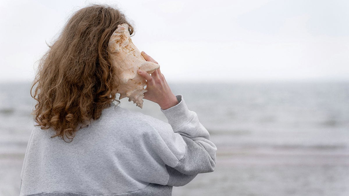 Person holding a large seashell to their ear while standing by the sea, symbolizing The Creative Guide Expressions listening playlists that spark reflection, quiet attention, and creative thinking.