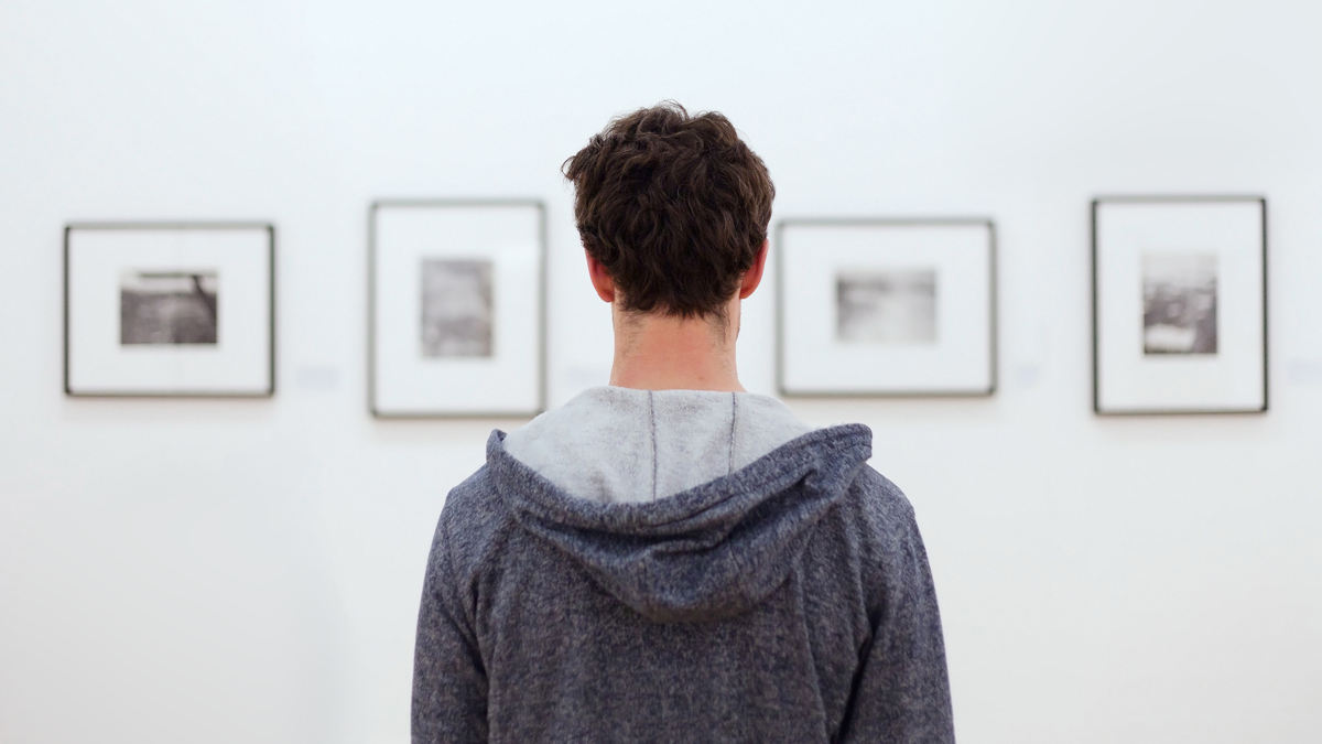 Person standing in front of framed black and white photographs in a gallery, symbolizing The Creative Guide’s photography gallery with images that include full camera settings as a learning tool.