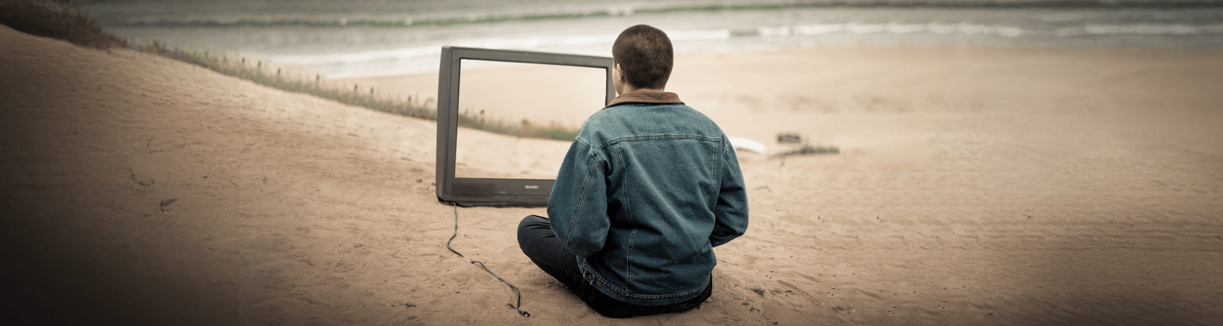 A person in a denim jacket sits on a sandy beach facing an old television screen with no image, the ocean waves visible beyond the frame. The surreal scene suggests reflection, perspective, and altered ways of seeing, aligning with The Creative Guide Podcast themes of Expressions courses on creativity and communication, followed by photography practice and insight.