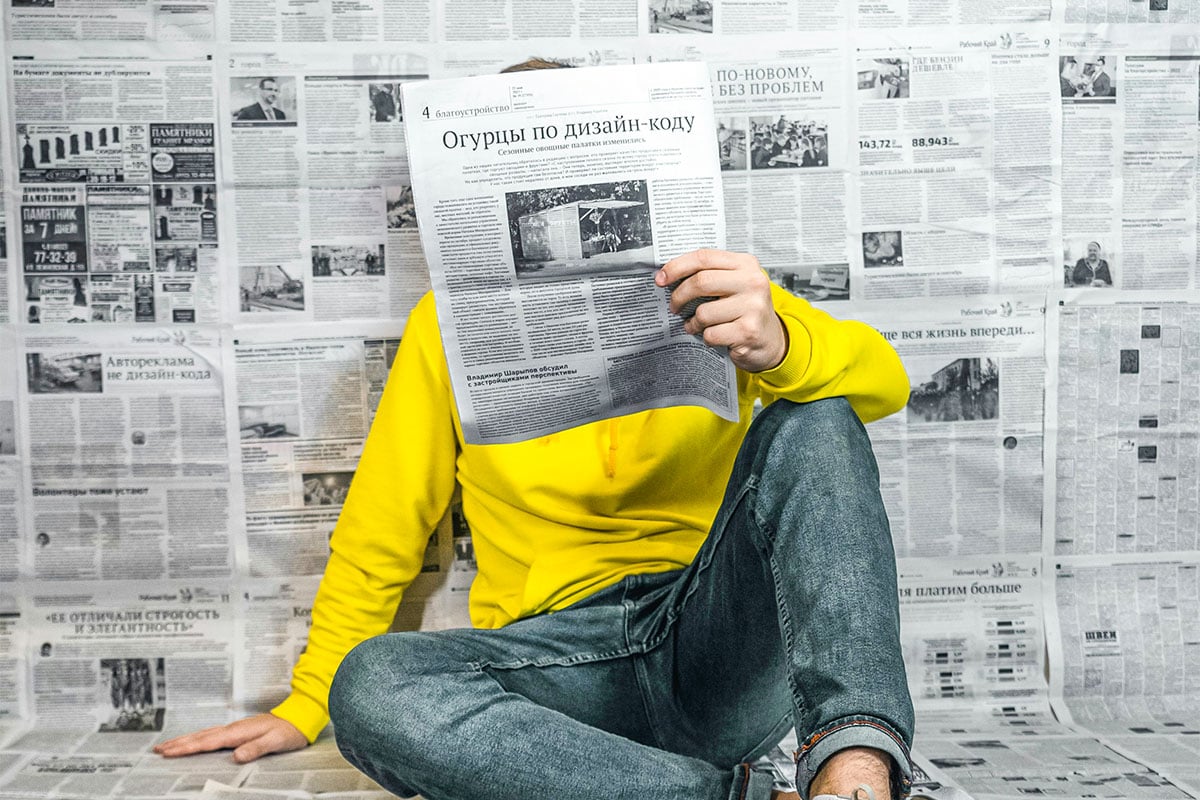 Person in a bright yellow hoodie sitting cross-legged and reading a newspaper, with a wall of newspapers behind them. Conceptual image used by The Creative Guide to represent the newsletter, highlighting news, updates, and creative insights.