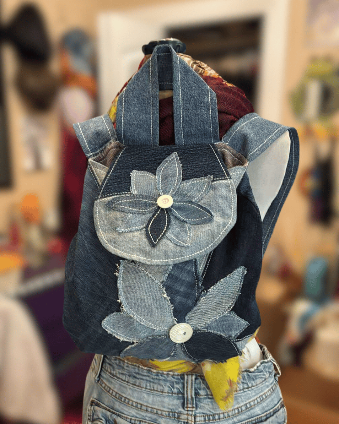 Handmade upcycled denim backpack bag created from recycled jeans and decorated with dimensional denim flower appliqués. This boho patchwork backpack purse features layered blue jean textures, raw edge detailing, sustainable fashion elements, and artistic textile embellishments.