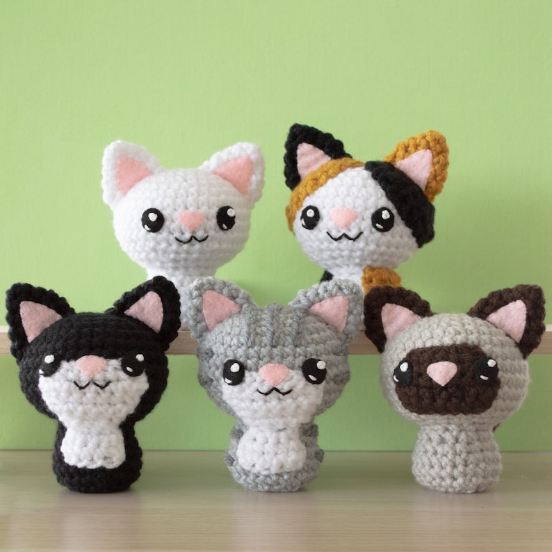 Five crochet amigurumi cats on light wood surface with green background — white cat, calico, tuxedo, gray tabby, and Siamese.