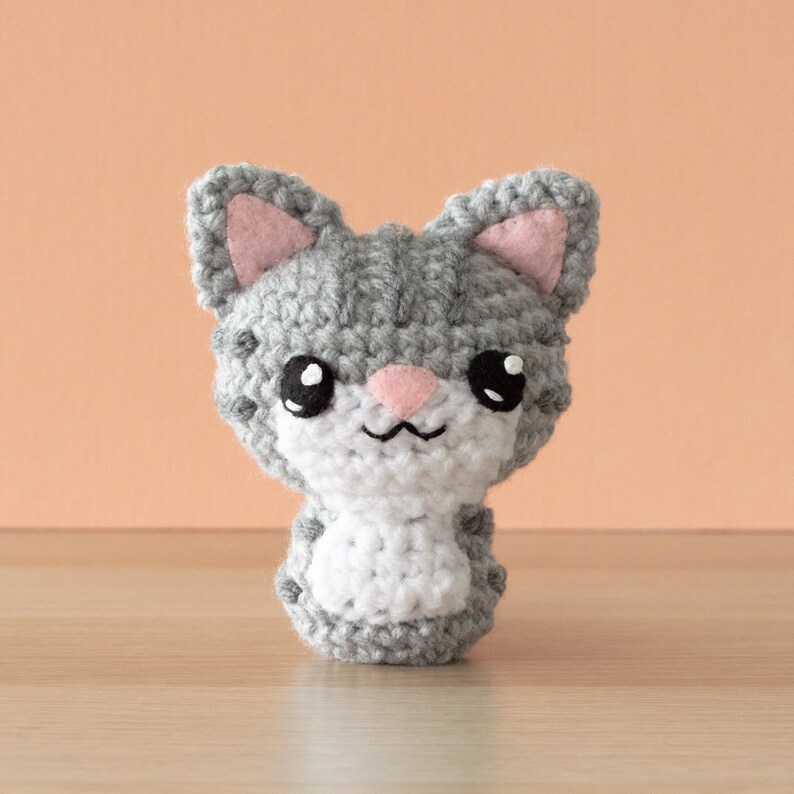 Crochet amigurumi gray tabby cat with salmon wall and light wood surface.