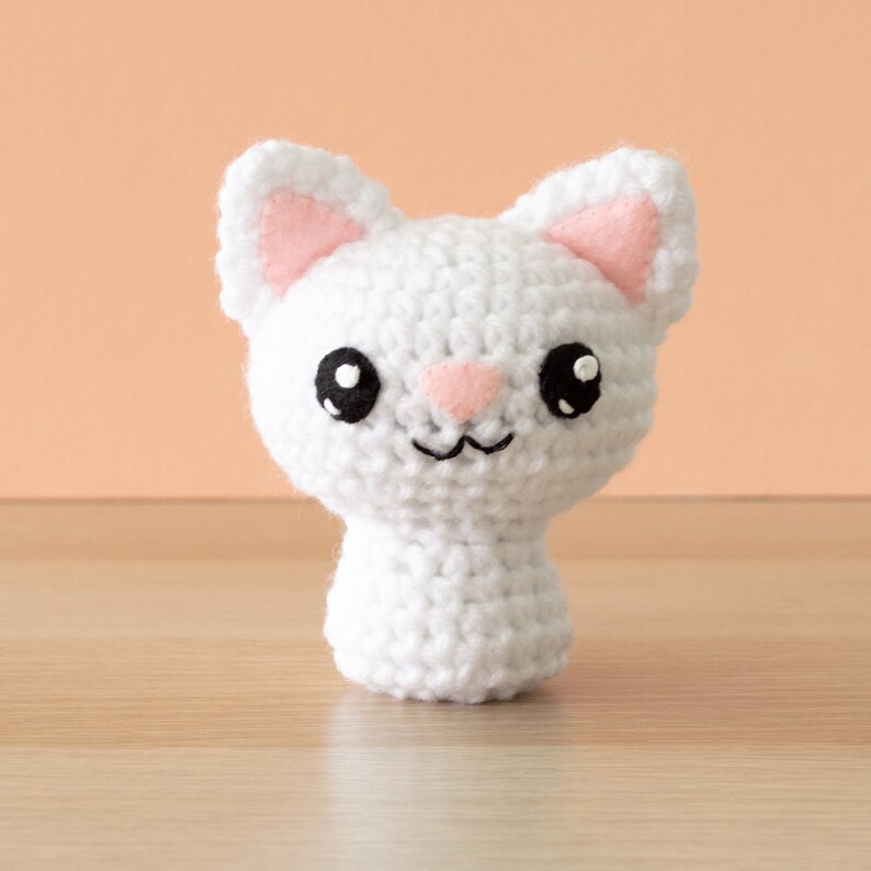 Crochet amigurumi white cat with salmon wall and light wood surface.