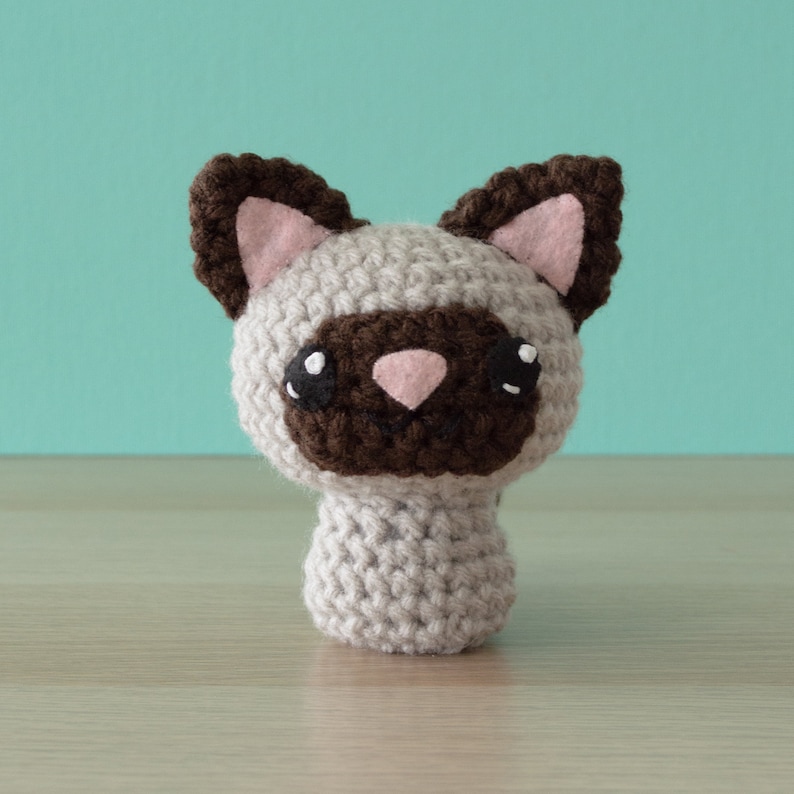 Crochet amigurumi Siamese cat with aqua wall and light wood surface.