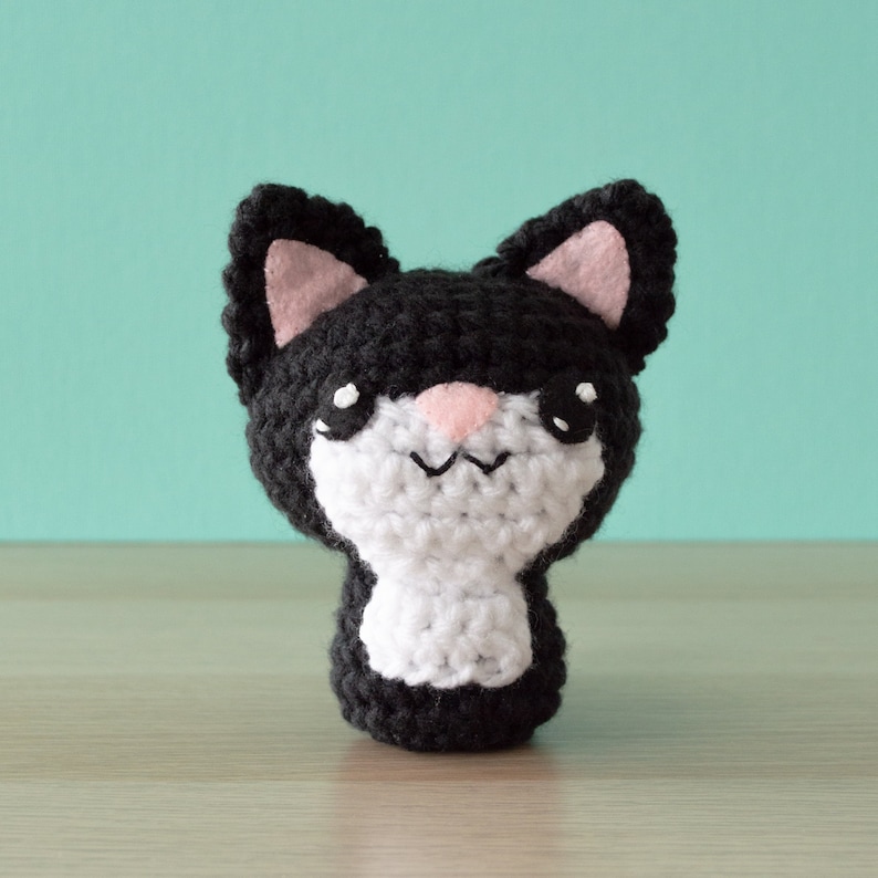 Crochet amigurumi black tuxedo cat with aqua wall and light wood surface.