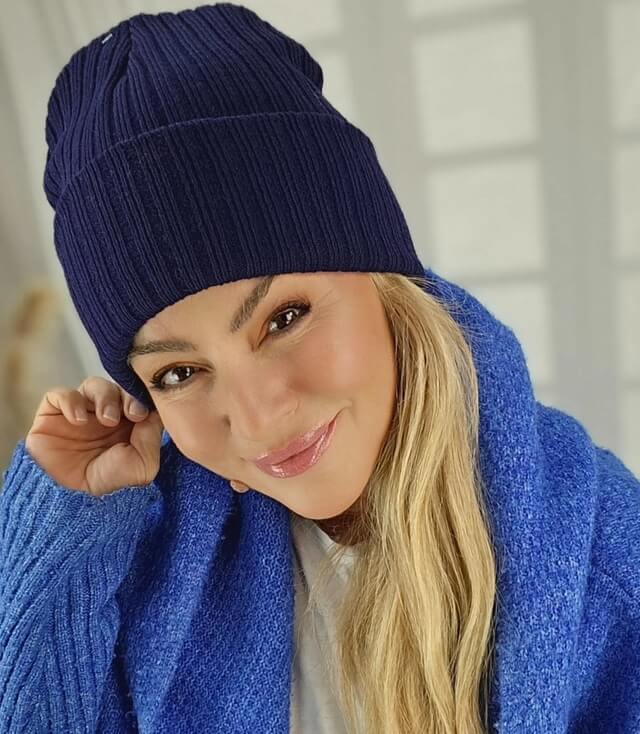 Frau mit langen blonden Haaren, die eine blaue Mütze trägt und in eine blaue Strickjacke gehüllt ist.
