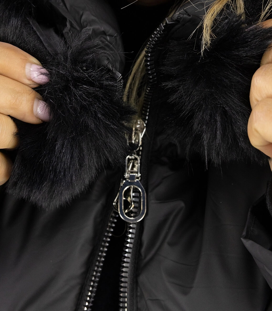 Detailansicht der schwarzen Jacke mit Kunstfellkragen und silberner Zipper-Ziehung.