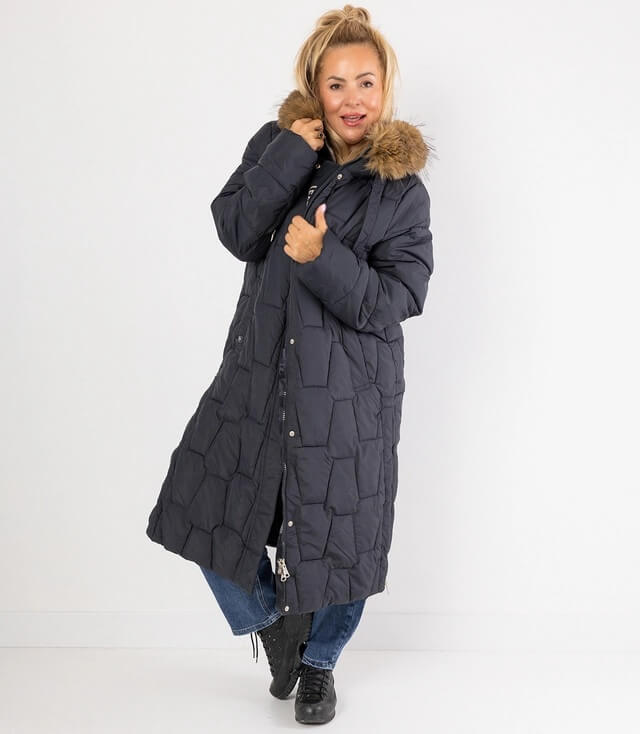 Dunkelblaue Steppjacke mit Kapuze und Kunstfellkragen, getragen von einer Frau in lässiger Pose.