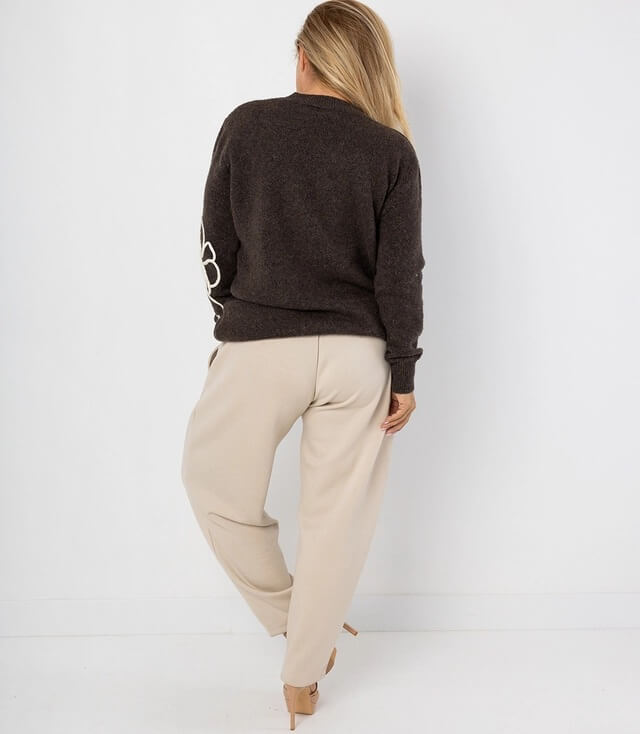 Dame in beiger Hose und grauem Pullover mit floralem Muster auf dem Arm, Rückenansicht.