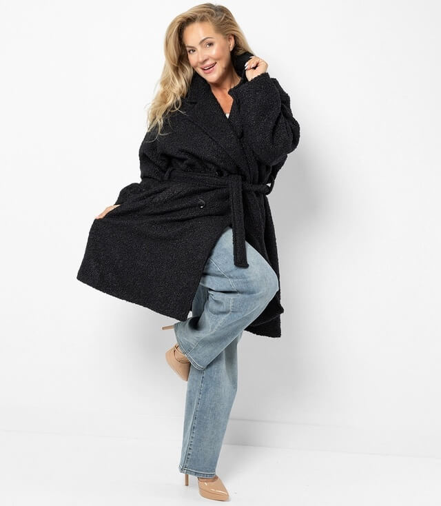 Gemütlicher oversized schwarzer Mantel mit Gürtel, getragen von einer Frau in Denim-Hose und hohen Schuhen.
