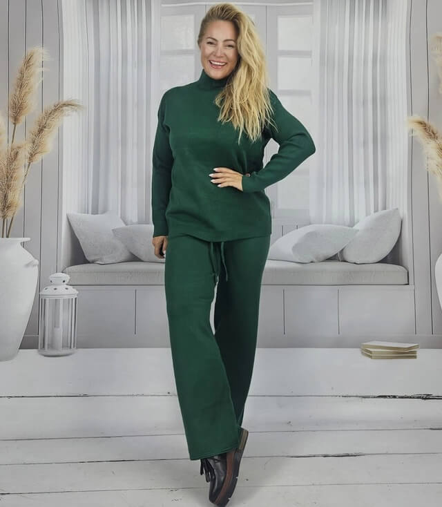 Frau in grünem Stoffanzug mit hohem Kragen, posierend vor heller Wohnzimmerkulisse.