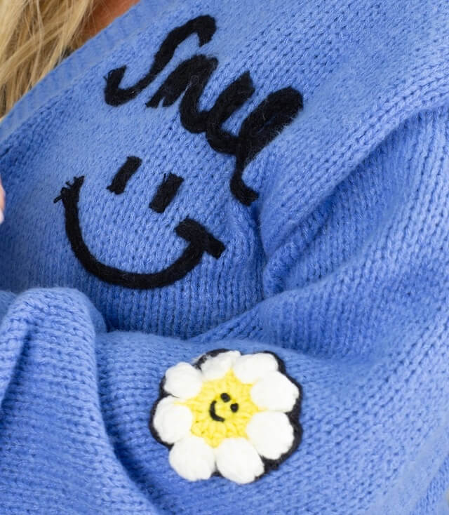 Blaue Strickjacke mit Smiley-Applikation und Blumendetail auf dem Ärmel.