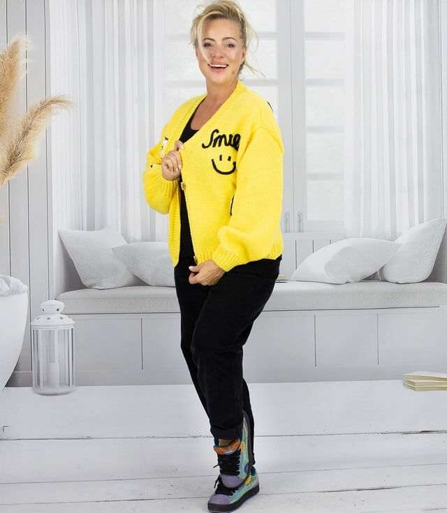Frau trägt eine gelbe Strickjacke mit Smiley-Print und schwarze Jogginghose in einem hellen Raum.