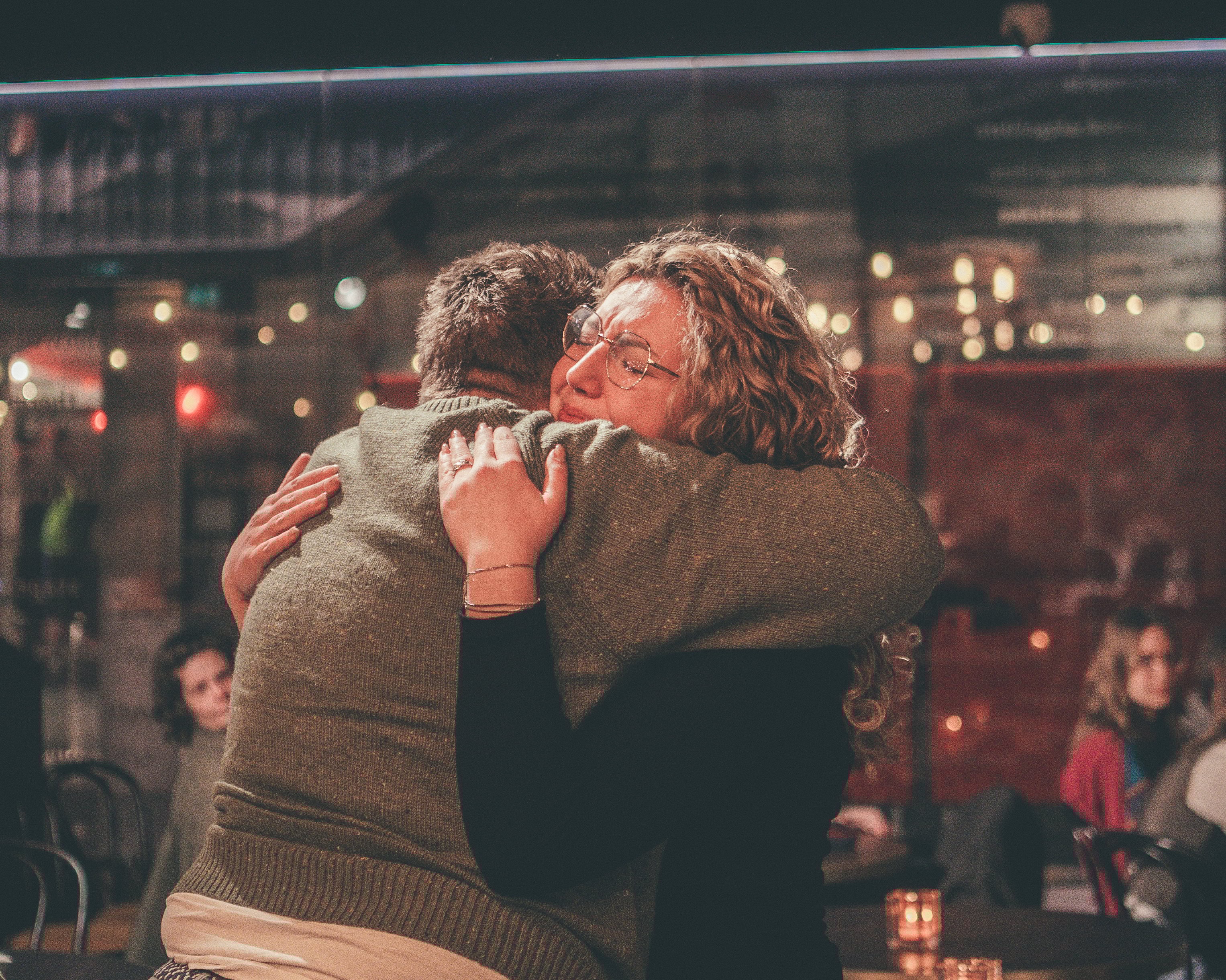 Twee mensen omhelzen elkaar in een warme, sfeervolle binnenruimte. Op de achtergrond zitten andere mensen aan tafels, met gedimd licht en kleine lampjes die zorgen voor een intieme setting.
