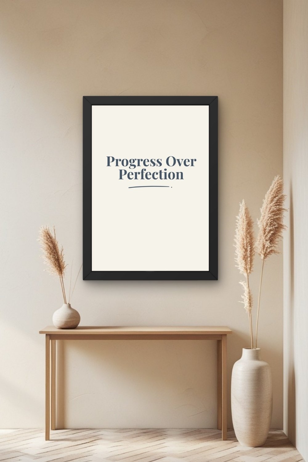 A navy typographic wall art print in a black frame on a warm neutral wall above a thin wooden console table with a round ceramic vase of pampas grass on one side and a tall vase of pampas grass on the other, on a herringbone wood floor.
