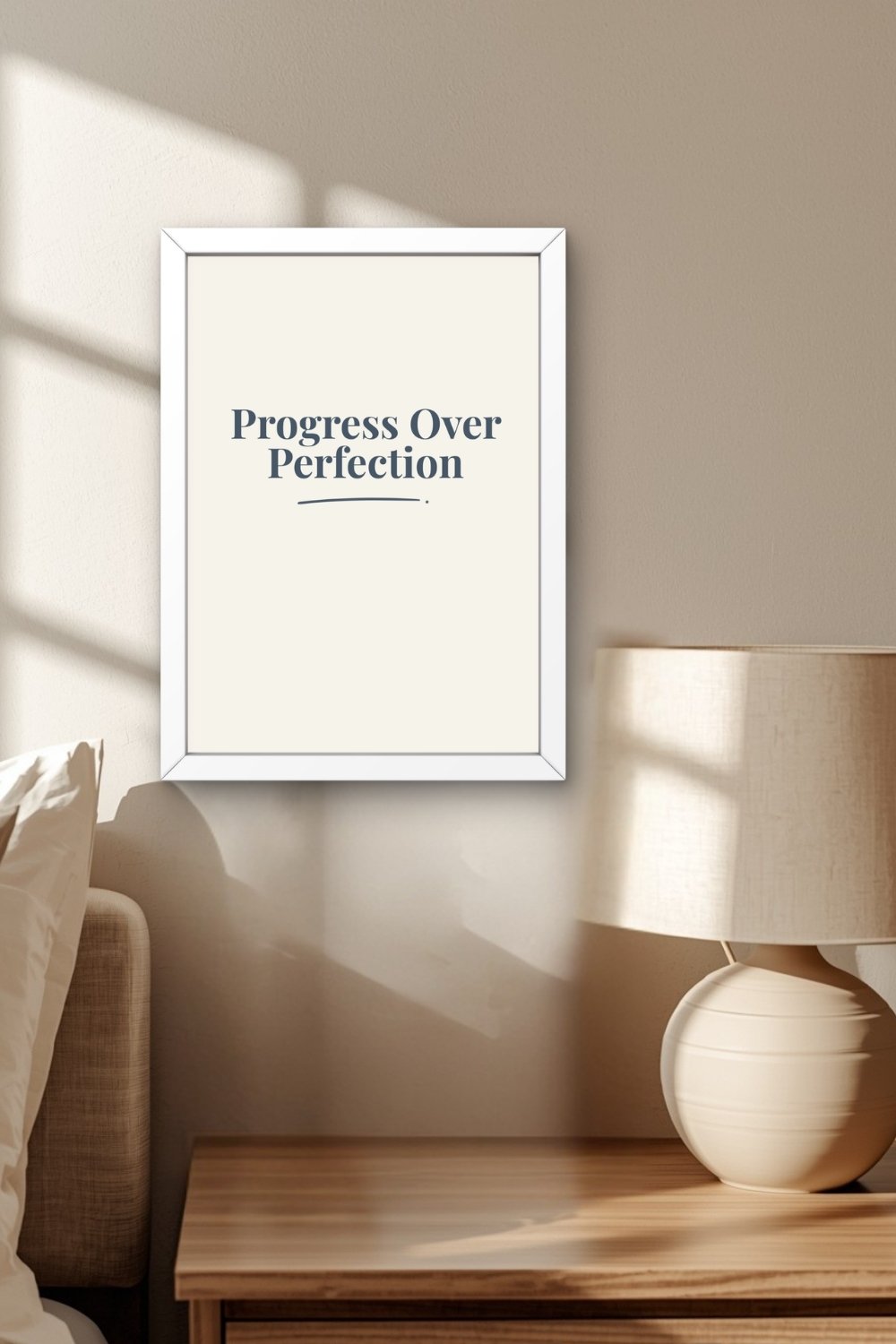 A navy typographic wall art print in a white frame on a warm neutral wall above a wooden nightstand with a round ceramic table lamp beside a linen upholstered headboard in a calm, softly lit bedroom.