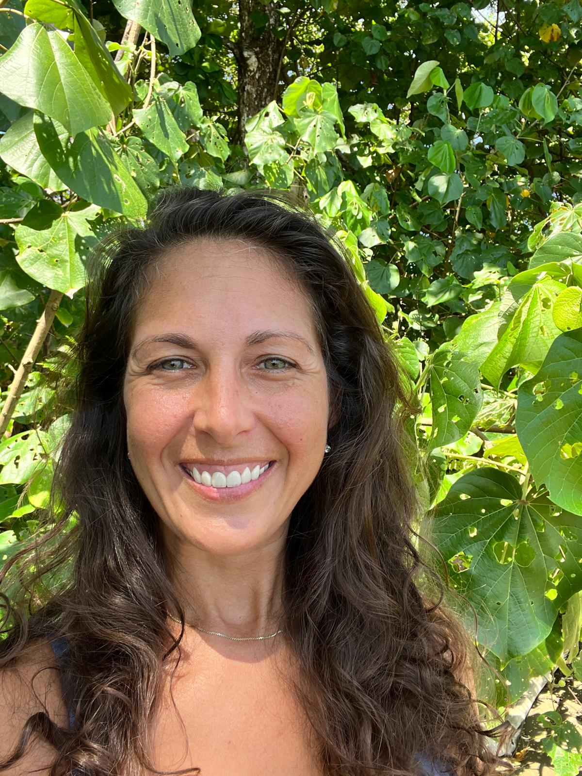 A dark-haired woman with green eyes smiles brightly in the green jungle.