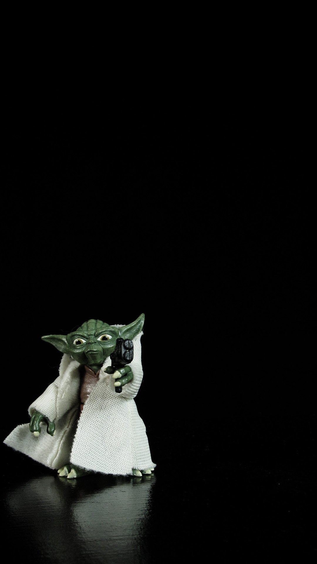 Yoda action figure on a black  background. He is pointing a blaster at the viewer.