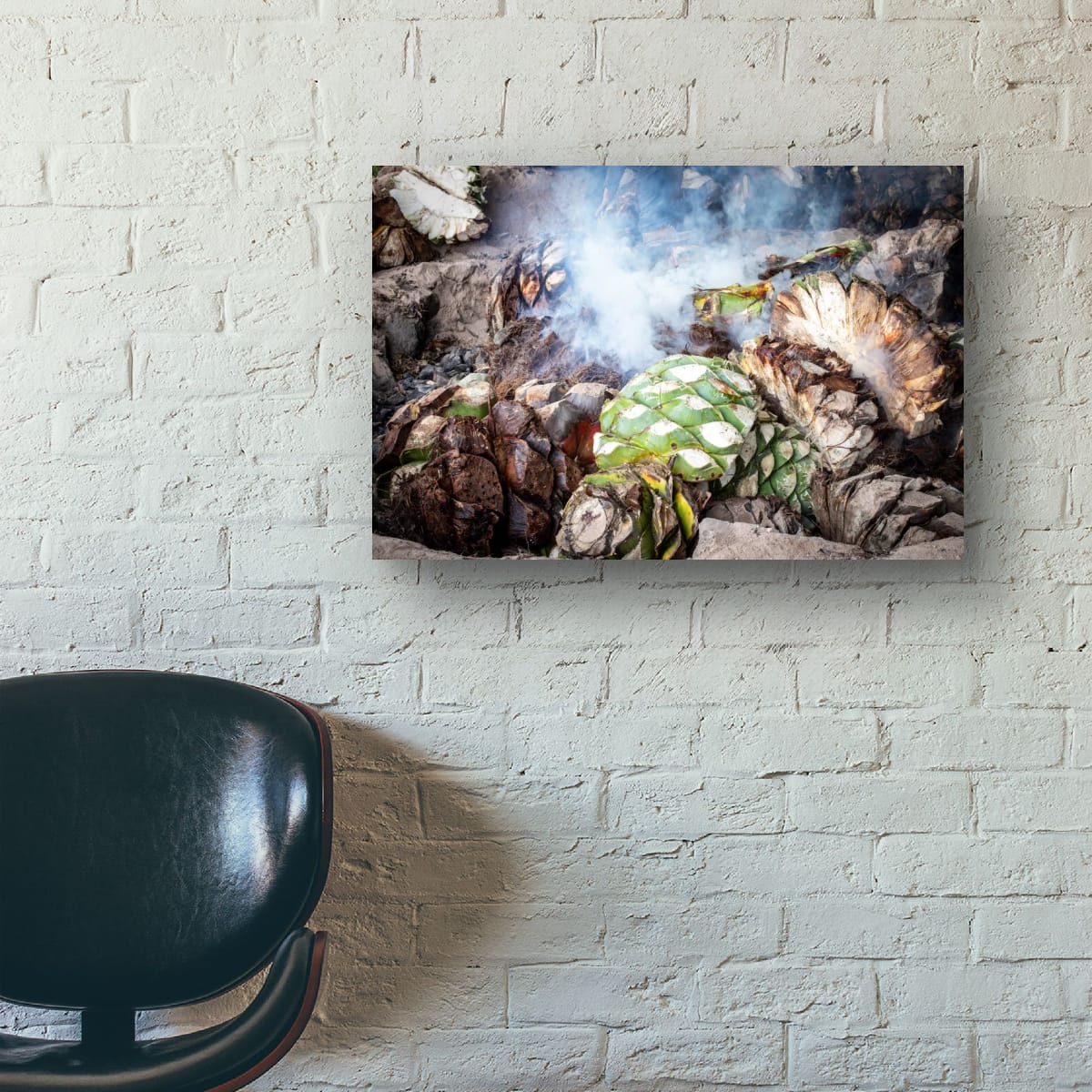A large horizontal print of a mezcal agave roasting scene hangs on a painted white brick wall above a black leather office chair.