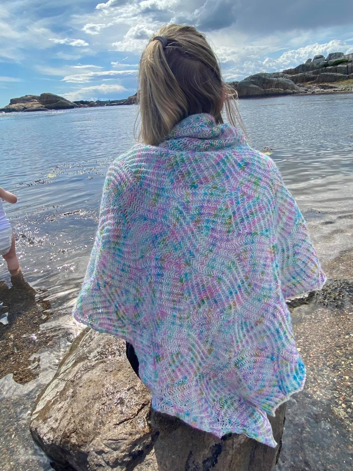 A woman is wearing a pastel/neon coloured Waterfall Shawl, sitting on a stone on a beach