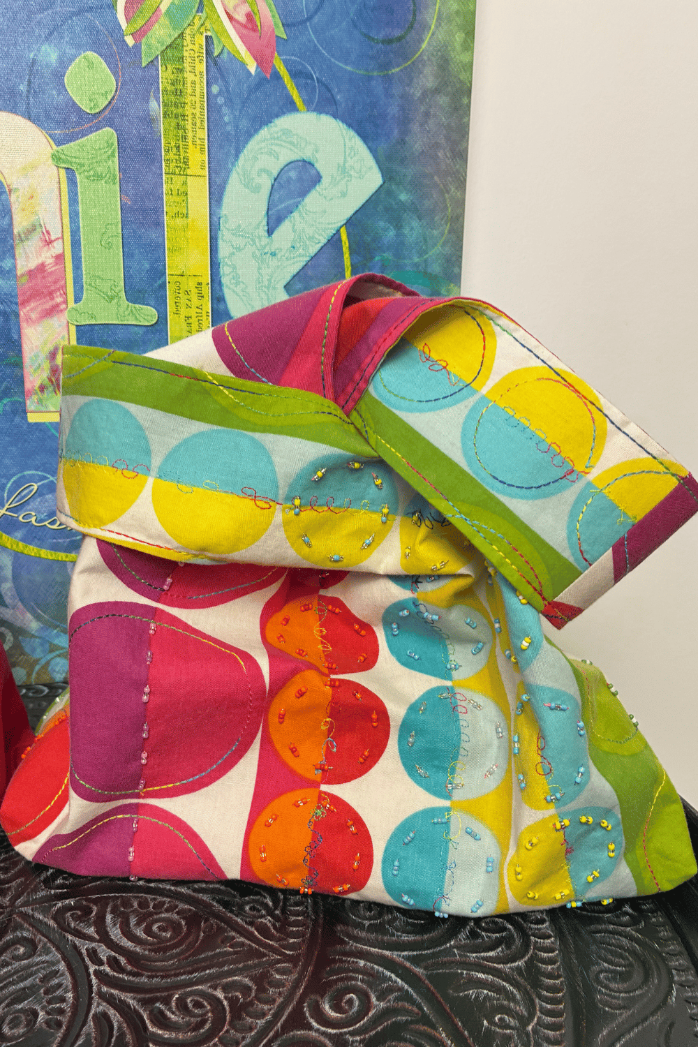 Front view of a handmade upcycled knot bag made from colourful circle print fabric with decorative rainbow stitching, bead embellishments, and a soft sculptural shape.