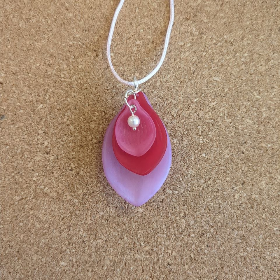 A pendant made of three stacked petal shapes in different shades of pink and red. a pearl detail sits at the top of the innermost petal. The petals are connected with silver toned metal rings and strung on a pale pink cord.