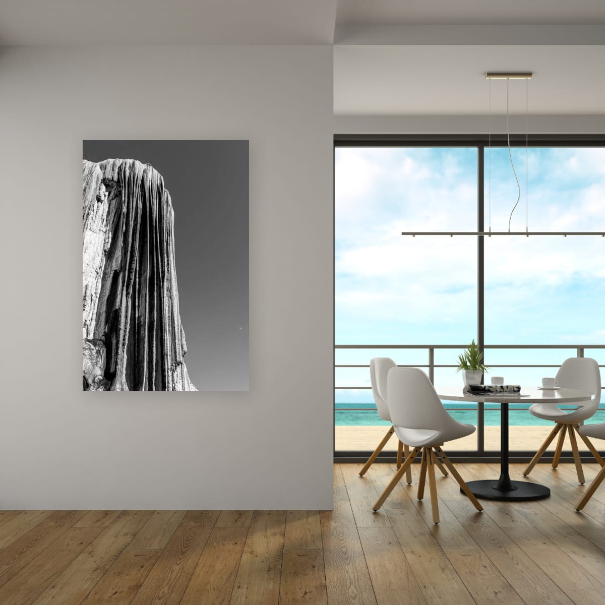 A large vertical canvas print of the black and white Hierve el Agua formation hangs on a white wall in a modern, light-filled dining space featuring hardwood floors and large glass doors opening to a coastal view.