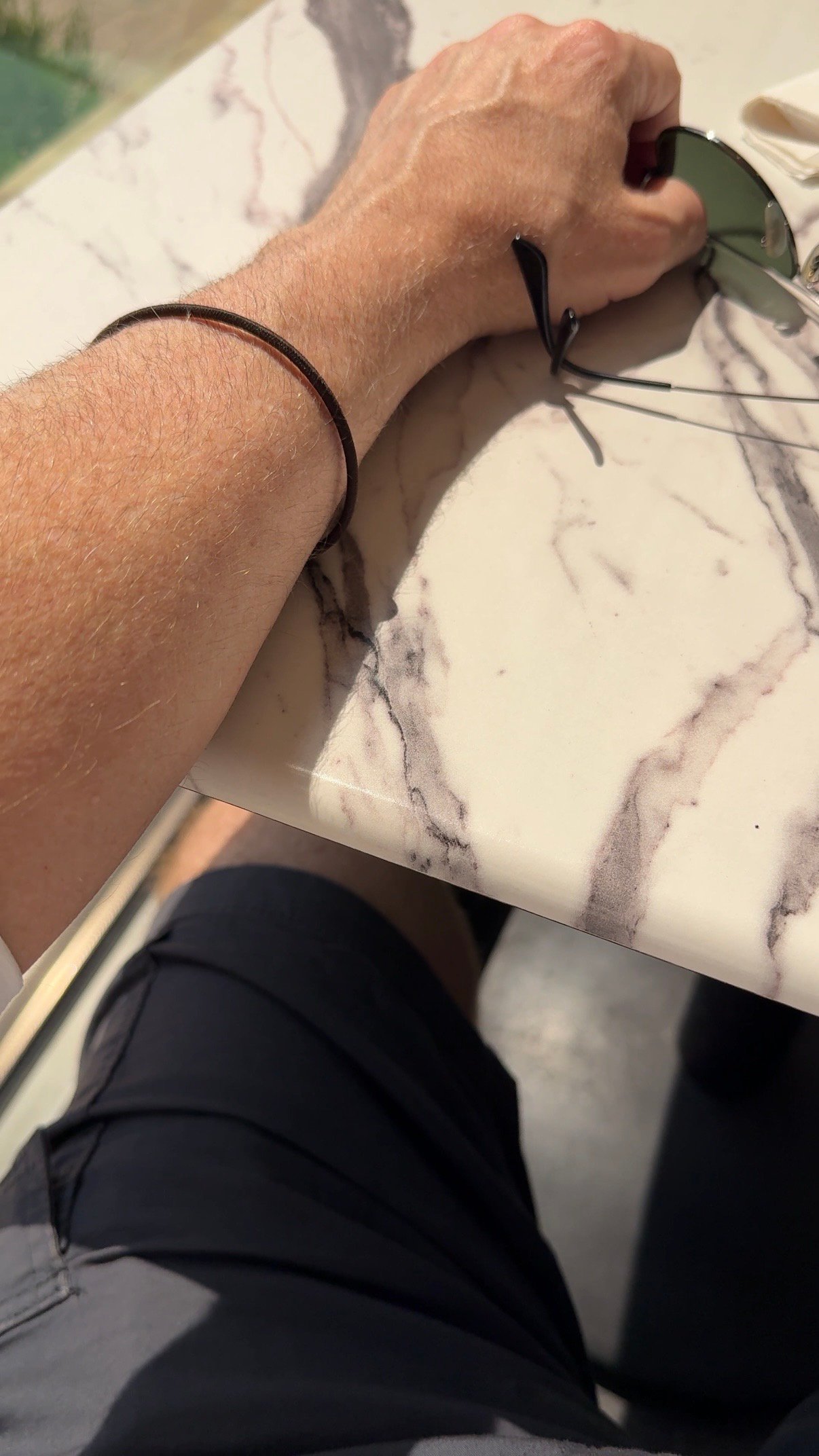 Close-up editorial photograph of a sunlit forearm resting on a marble café table beside sunglasses, capturing a quiet moment of masculine daily ritual and natural light.