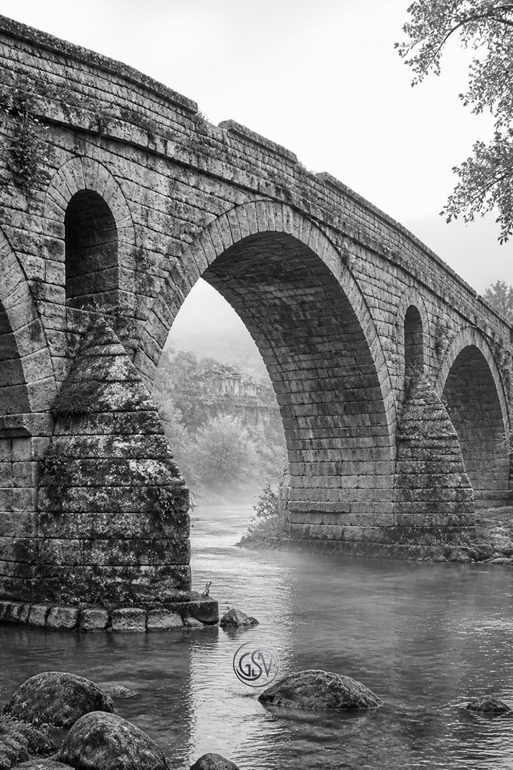 Grayscale coloring page of an ancient Roman stone arch bridge over a river with weathered stone and mist