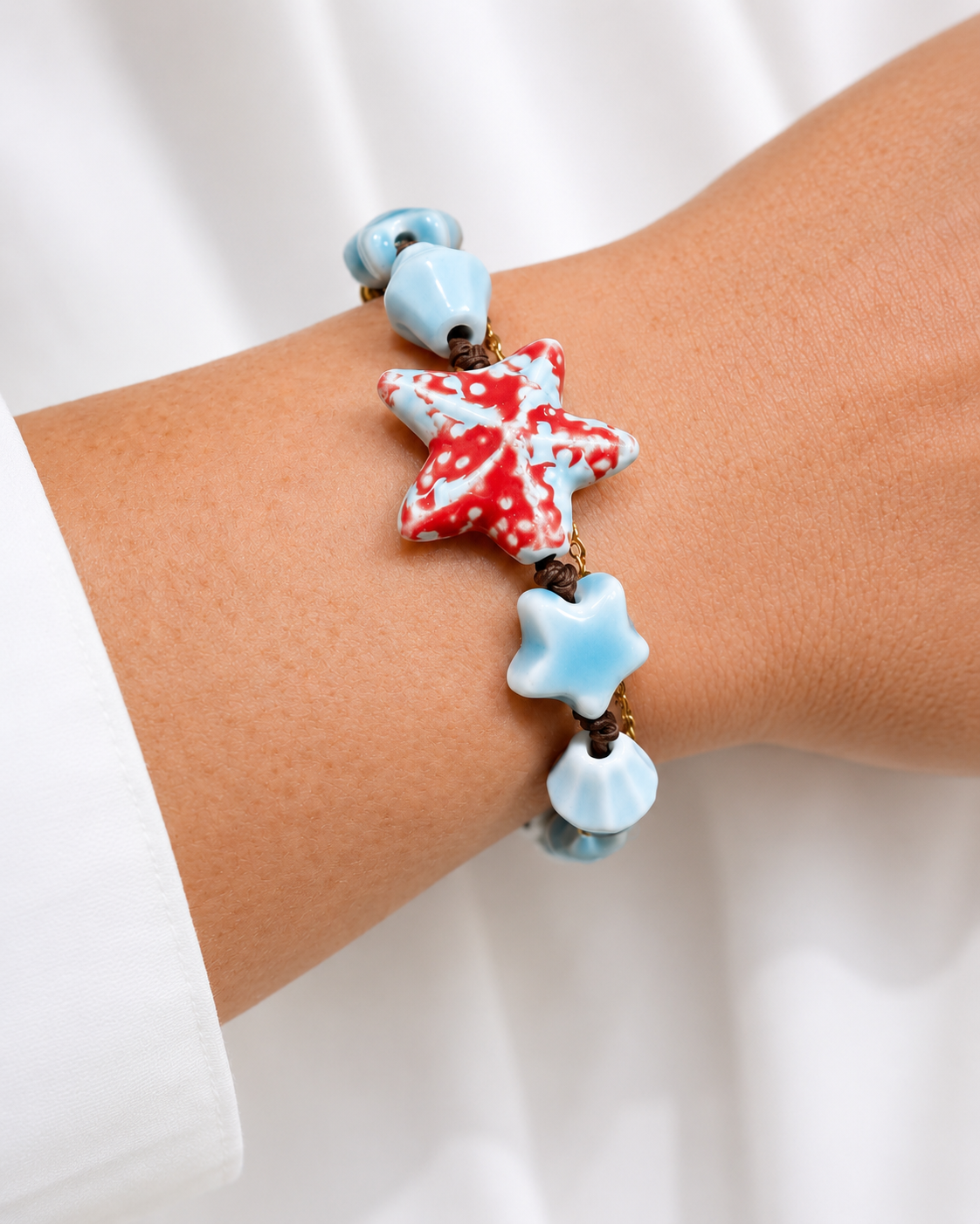 Bracciale artigianale ispirato al mare con stella marina e conchiglia in ceramica smaltata, indossato al polso su sfondo chiaro.