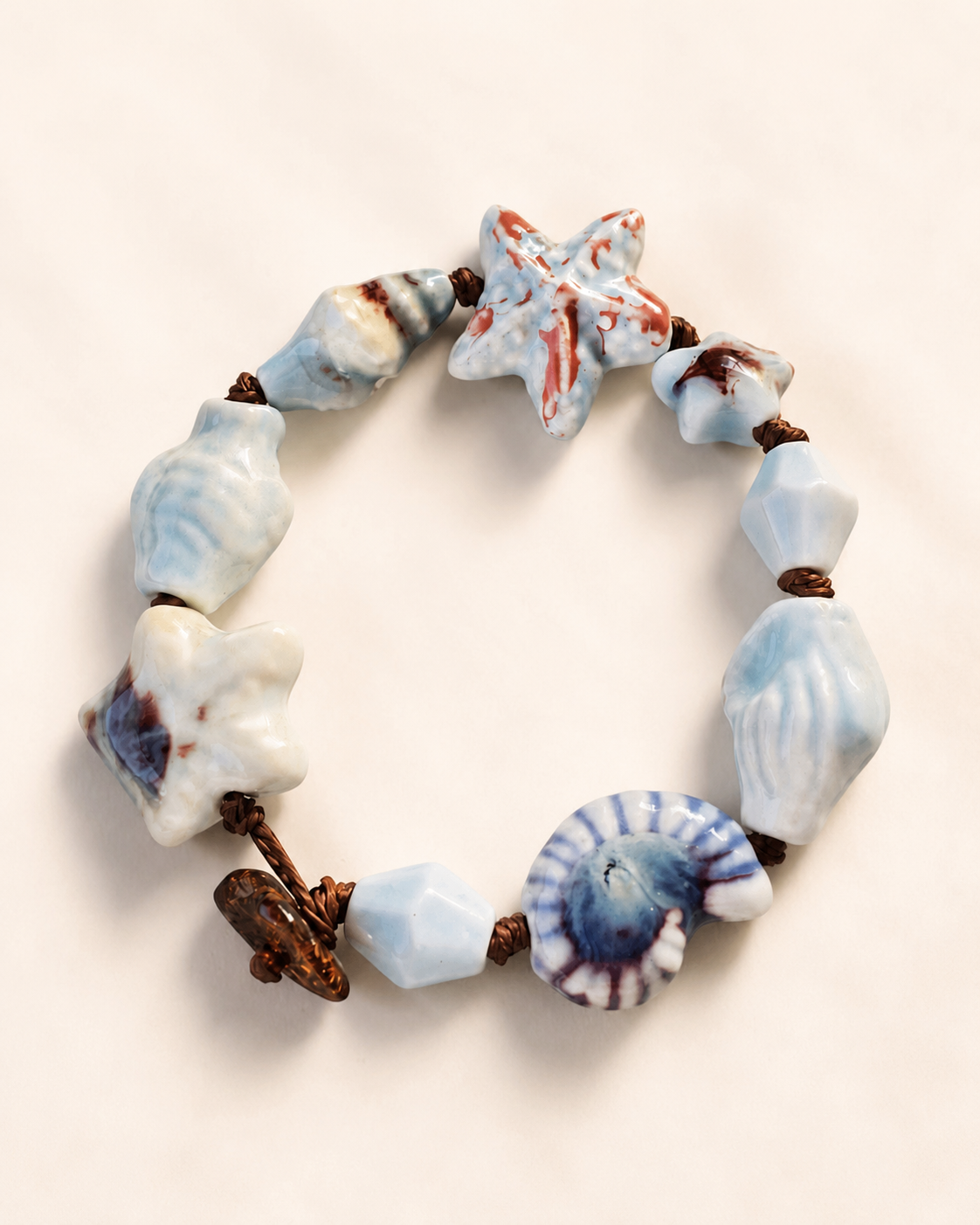 Dettaglio del bracciale artigianale con stella marina e conchiglia in ceramica smaltata, cordoncino marrone e componenti in acciaio inox.