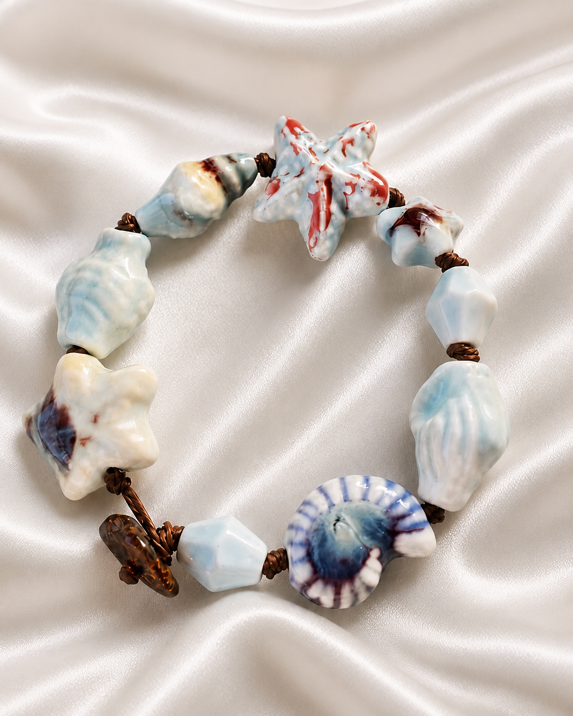 Bracciale artigianale con elementi in ceramica smaltata a forma di stella marina e conchiglia, fotografia su sfondo chiaro in stile catalogo.
