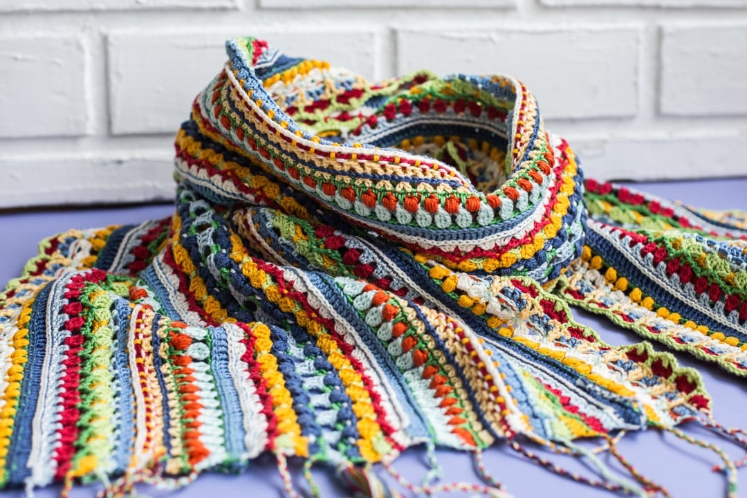 Colorful scrappy crochet sampler scarf Blossom made with fingering-weight cotton yarn, featuring a variety of textured crochet stitches, bright primary colors, and fringe.