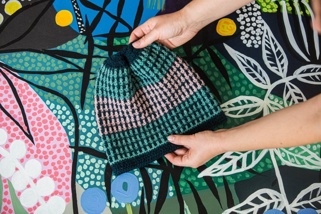 Colorful striped overlay mosaic crochet hat held in both hands against a bright background.