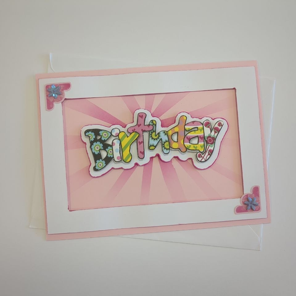 A landscape card in pink with a white frame. The word "Birthday" in multiple colours and patterns of bubble text floats over a pink blended starburst effect. Two small iridescent corner stickers sit in opposite corners of the frame.