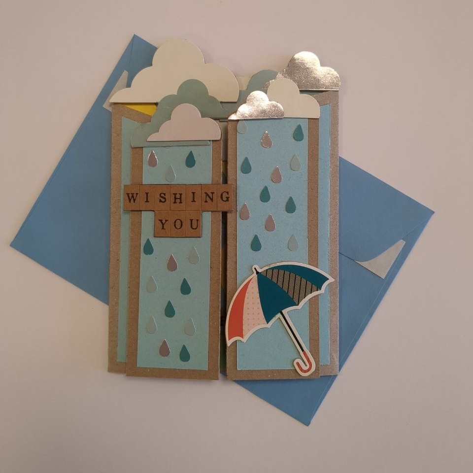 A closed concertina folded card with fluffy white, blue and silver clouds. Blue panels on craft card feature falling raindrops, a partial sentiment "wishing you", and an umbrella.