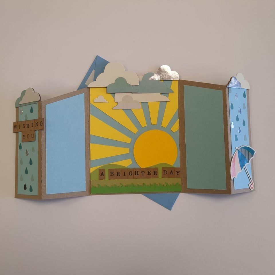 A concertina folded card, open to show the inner image of a sun bursting over hills under fluffy clouds, with the partial sentiment "a brighter day". Blue panels at the side are blank