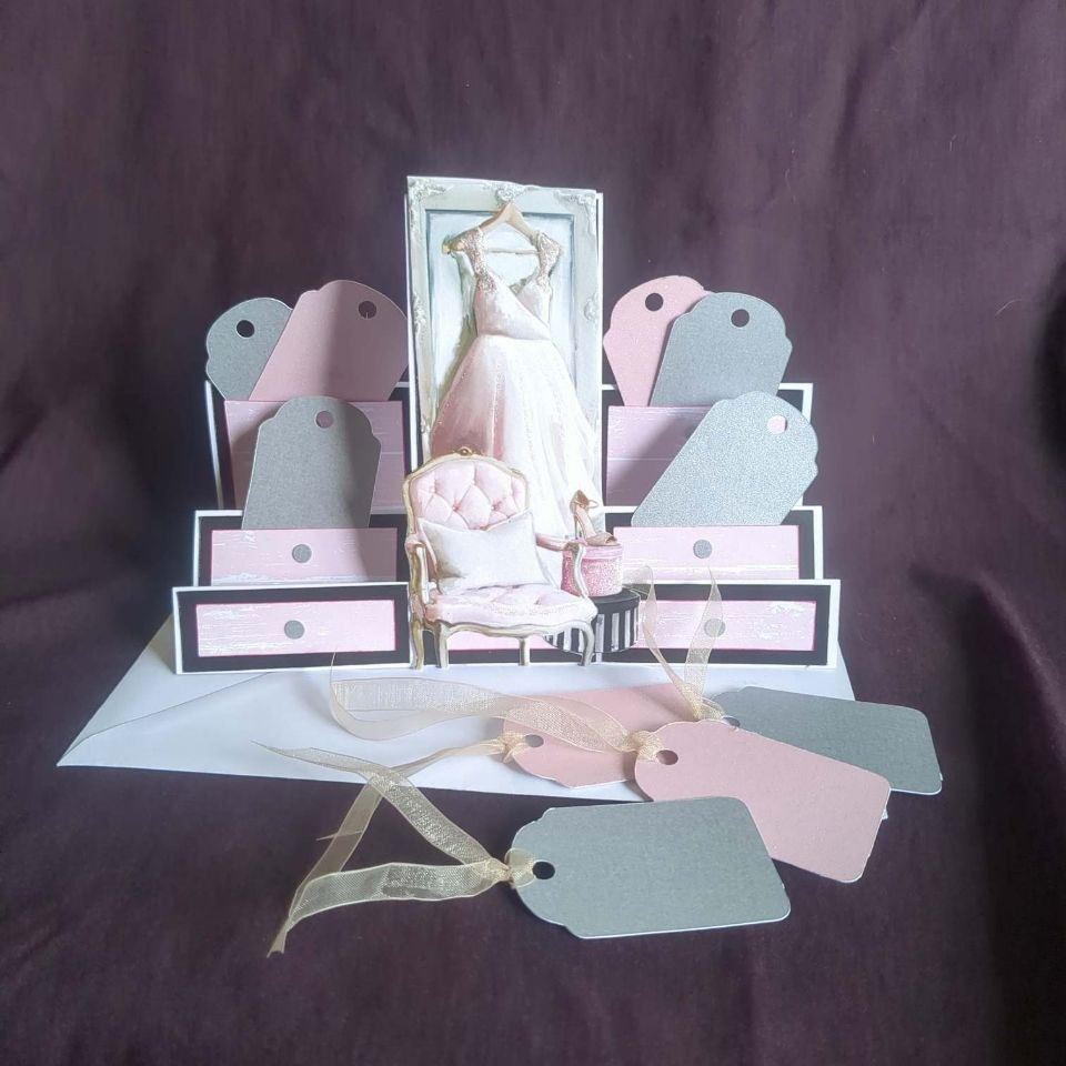 A stepper card with lower sides and a higher middle. The middle panel features an image of a pink chiffon prom dress hanging on a door, in front of which are a padded armchair and hat boxes. The steps of the card have been made to resemble drawers, and gi