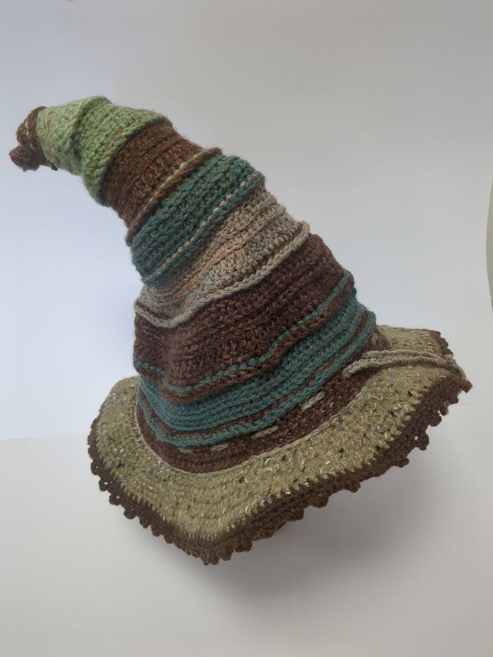 A witchy hat with a curled point. The hat is crocheted in stripes of natural greens and browns, with a picot edge around the brim and a drawstring.